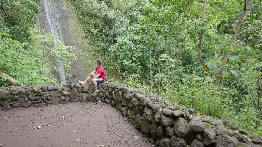 A female photographer captures a shot of a man relaxing on a stone wall while viewing a beautiful waterfall in the jungle