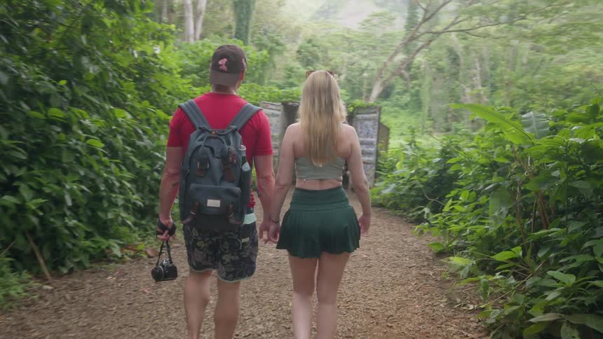 A young couple holds hands while walking down a trail through a lush tropical forest