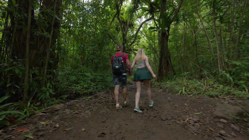 A young couple holds hands while hiking on a muddy trail through a dense, lush tropical rainforest