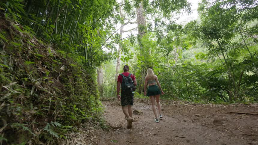 A couple hikes up a muddy trail through a lush, tropical bamboo forest in slow motion