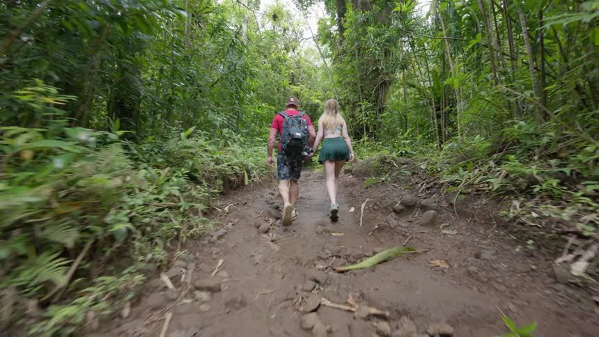 An adventurous couple holds hands while hiking along a muddy, rocky trail deep within a lush tropical jungle
