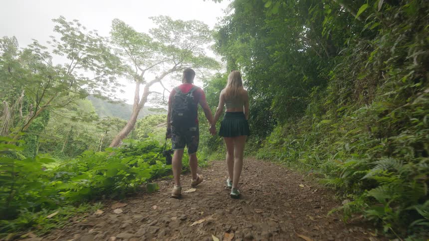 A couple holds hands while walking together down a lush green hiking trail in a tropical jungle