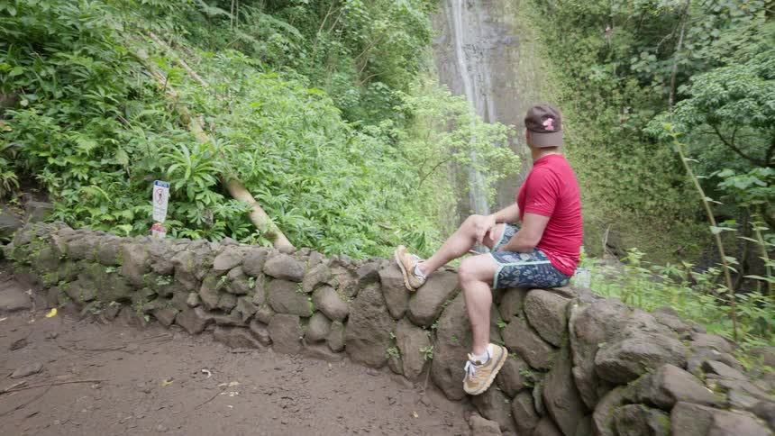 A young man in athletic wear sits on a rock wall admiring a scenic waterfall in a lush tropical jungle