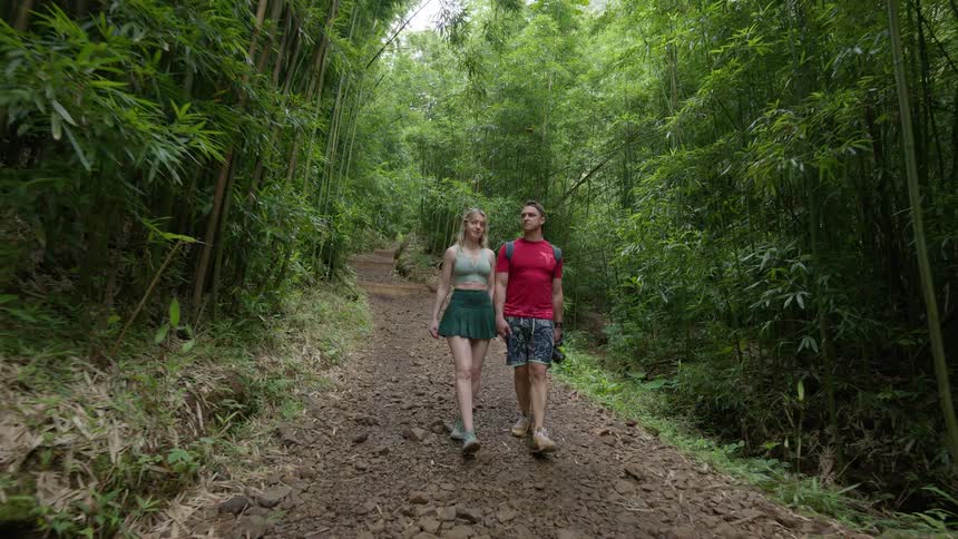 An adventurous young couple holds hands while walking along a scenic trail through a lush bamboo forest