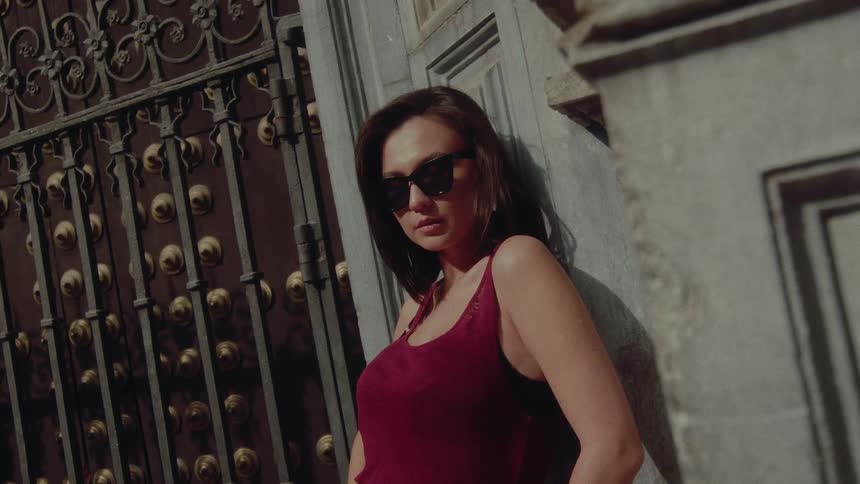 A beautiful brunette woman in sunglasses poses against a wall next to an old, ornate metal gate in the city