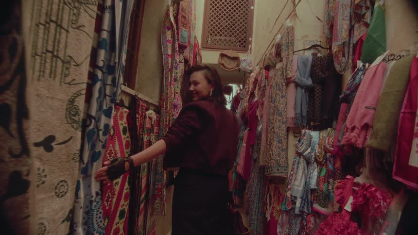 A fashionable woman smiles as she walks in slow motion through a narrow bazaar filled with colorful textiles and clothing