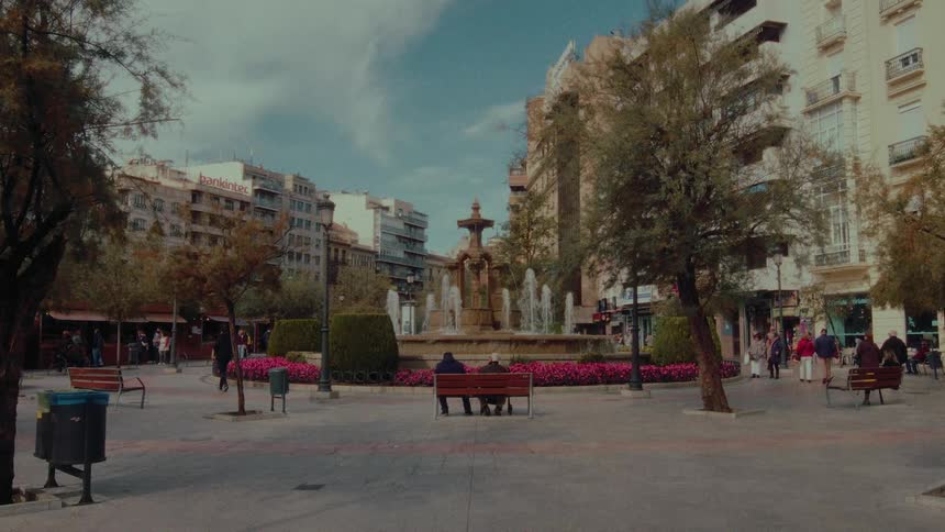 People relax on benches and stroll through a charming European city square with a grand fountain at its center