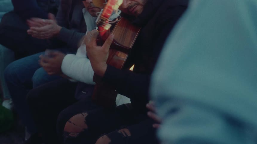 A musician plays an acoustic guitar while friends sit and clap along to the rhythm during a sunset gathering