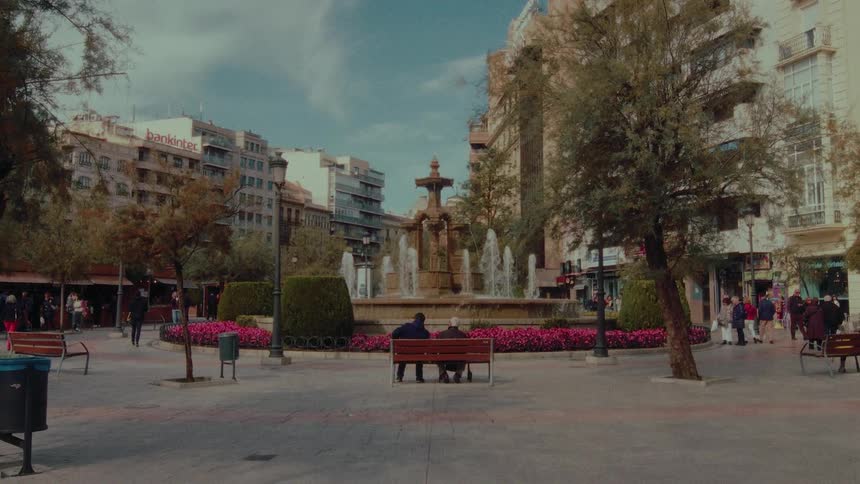 An elderly couple sits together on a park bench, sharing a quiet moment while watching a beautiful fountain in a bustling city plaza