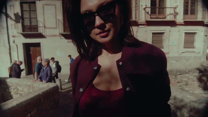 A chic woman wearing sunglasses and a maroon jacket breaks into a joyful smile on a sunny day in a historic European city