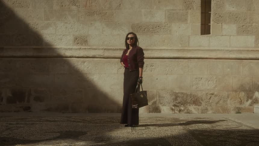A stylish woman in sunglasses stands in a sunbeam against an old stone building before looking at the camera