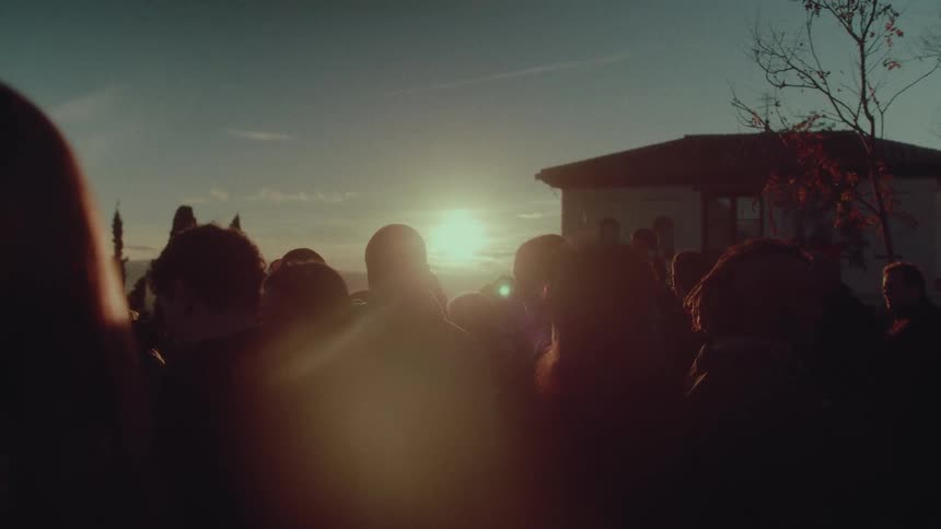 A crowd of people are silhouetted against the warm glow of the setting sun at an outdoor gathering