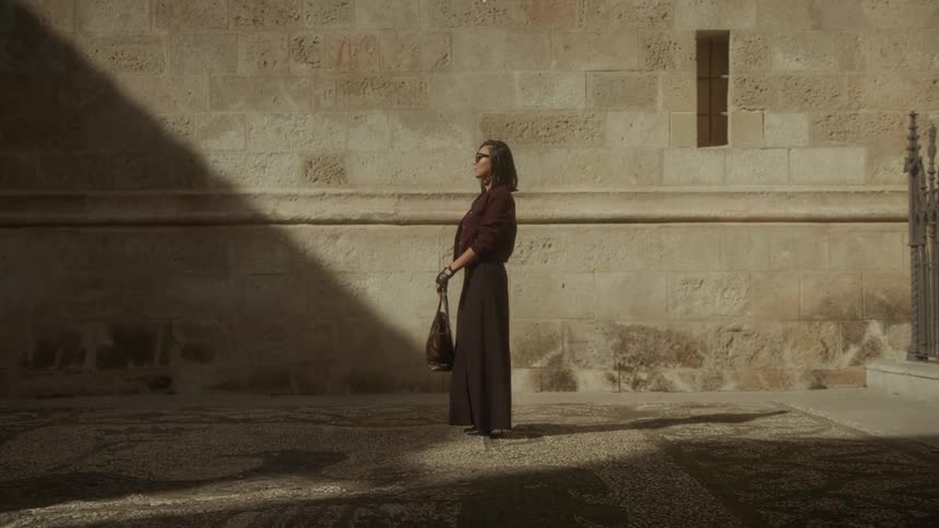 A stylish woman stands in a sunbeam against an old stone wall, looking up