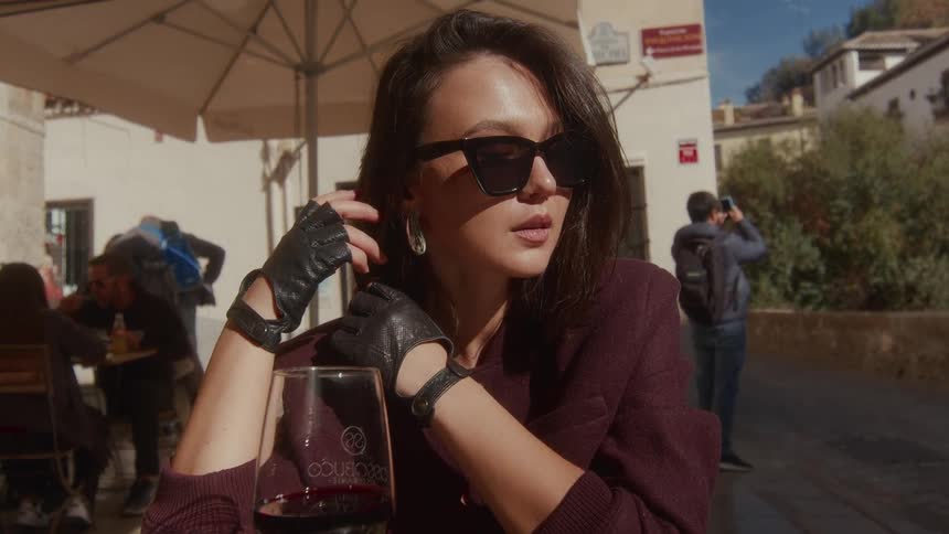 Close-up of a chic woman in sunglasses pensively sitting at an outdoor cafe table with a glass of red wine on a sunny day