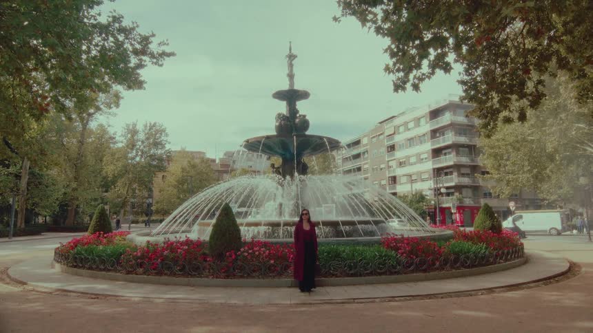 A woman in a long red coat stands serenely in front of an ornate, flowing water fountain in a city park