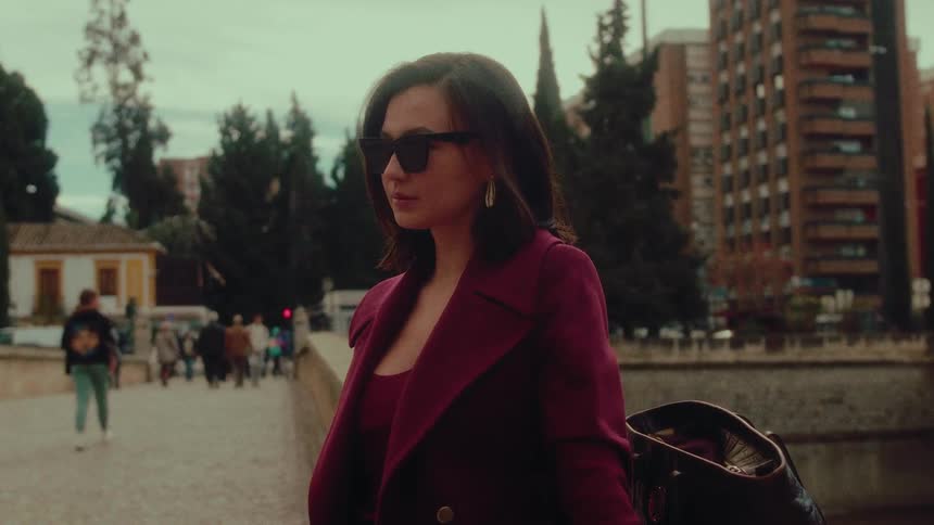 A stylish woman in a magenta coat and sunglasses stands thoughtfully on a bustling European city street