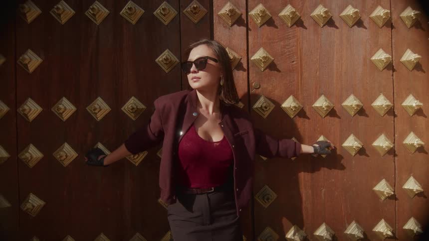 A stylish woman in sunglasses poses confidently against a large, ornate wooden door while soaking in the sunlight
