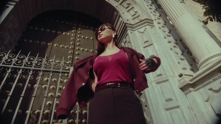 A cool and fashionable woman in sunglasses and gloves puts on her jacket in dramatic slow motion in front of an ornate, historic building