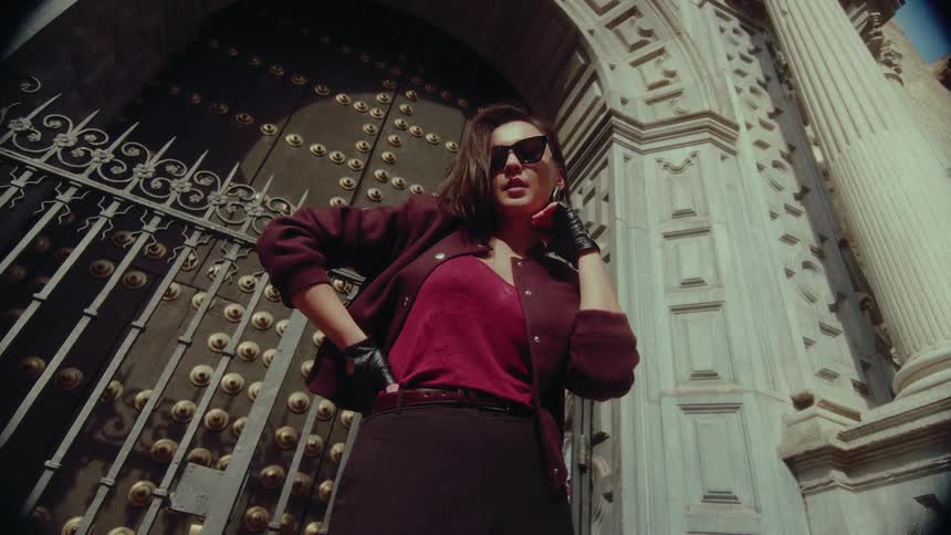 A stylish woman in sunglasses strikes a confident pose in front of a grand historic building, shot from a low angle for a powerful effect