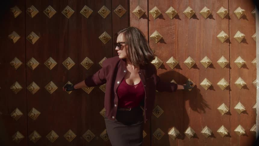 A stylish woman in sunglasses dramatically turns her head in slow motion while standing against an ornate, studded wooden door