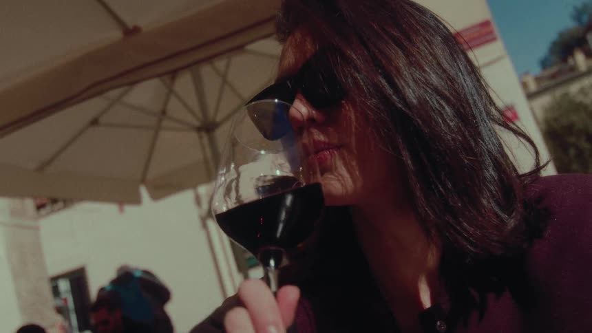 Sophisticated woman in sunglasses slowly sips red wine at a sun-drenched outdoor restaurant