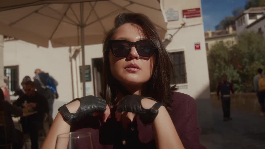A chic woman wearing sunglasses and leather gloves sits pensively at an outdoor cafe on a sunny day