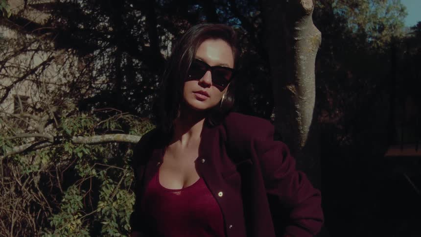 A stylish woman in sunglasses confidently poses in a sun-dappled garden on a sunny day