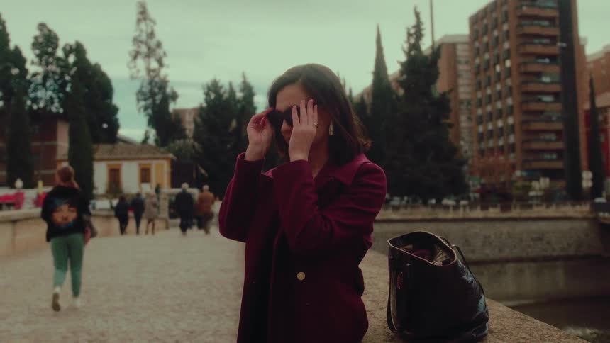 A stylish woman in a maroon coat puts on her sunglasses in slow motion while standing on a city bridge