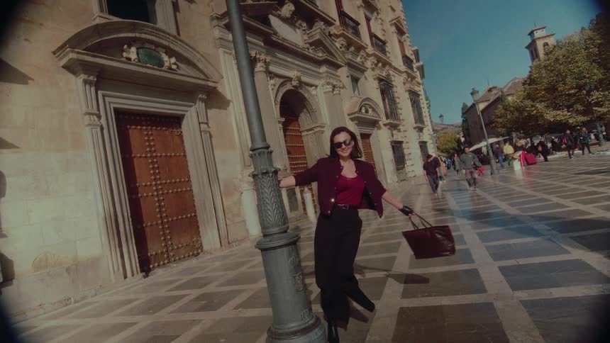 A stylish woman playfully spins around a lamppost in slow motion on a sunny day in a historic European city square