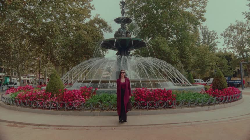 A woman in a stylish red coat stands still in front of a grand fountain in a city square on an overcast day