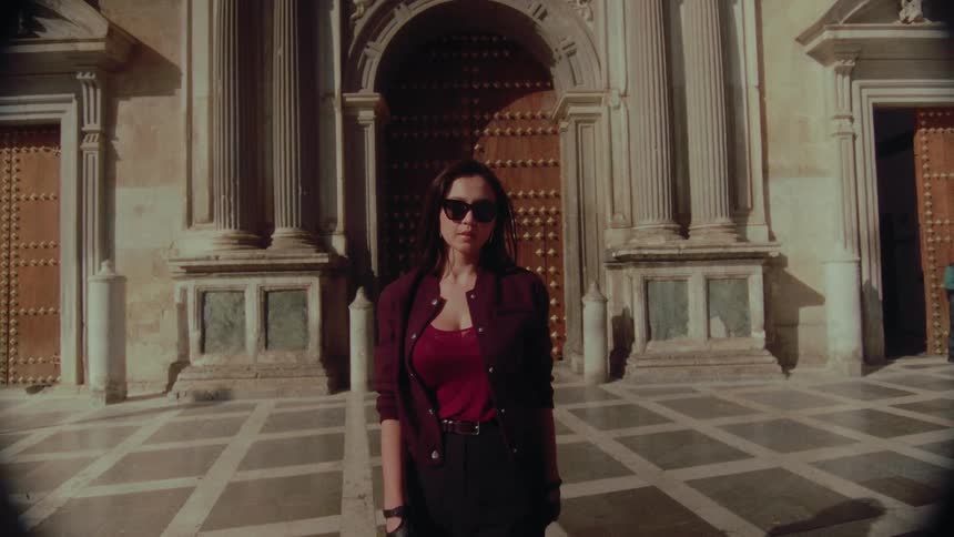 A mysterious woman in sunglasses and gloves walks in slow motion in front of an ornate historic building