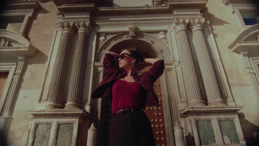 A mysterious woman with vintage style poses confidently in front of a majestic, ornate building on a sunny day