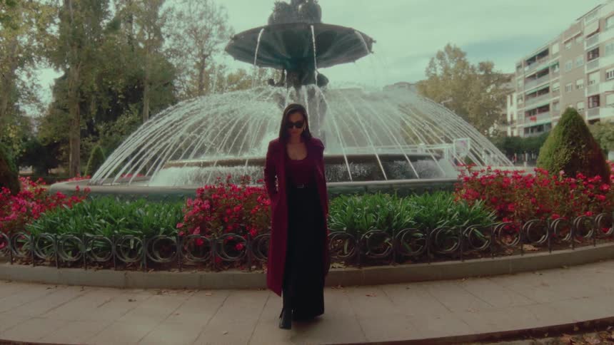 A chic woman in a maroon coat and sunglasses confidently walks towards the camera in front of an ornate city fountain