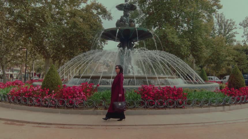 A stylish woman in a long maroon coat walks past an ornate water fountain in a beautiful city park