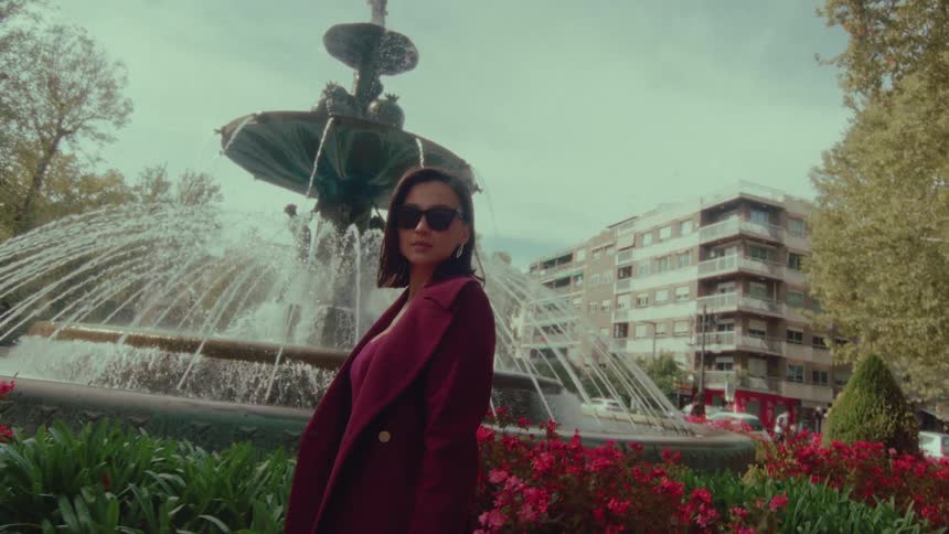 A stylish woman in a chic maroon coat and sunglasses poses confidently in slow motion by a beautiful city fountain