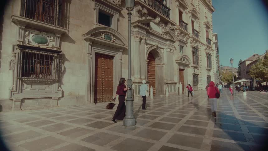 A stylish woman walks in slow motion through a sunny European city square and poses against a lamppost