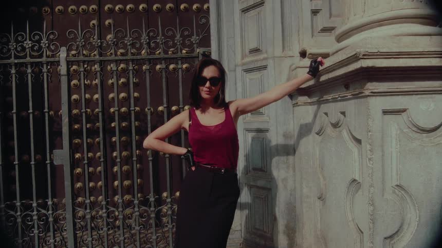 A stylish woman in sunglasses and black gloves poses confidently in front of a historic ornate gate on a sunny day