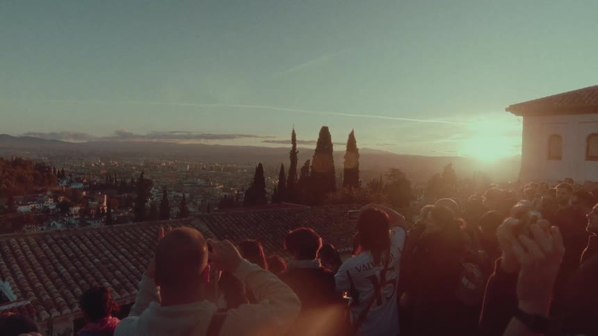 A large crowd of people gather at a scenic viewpoint, holding up their phones to capture the beautiful sunset over the city