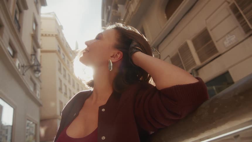 From a low angle, a woman smiles serenely as she soaks up the bright sun in a narrow city street