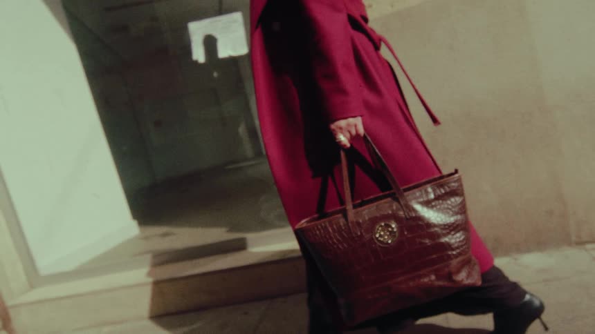 A stylish woman in a vibrant magenta coat carries a leather tote bag while walking in slow motion on a city street