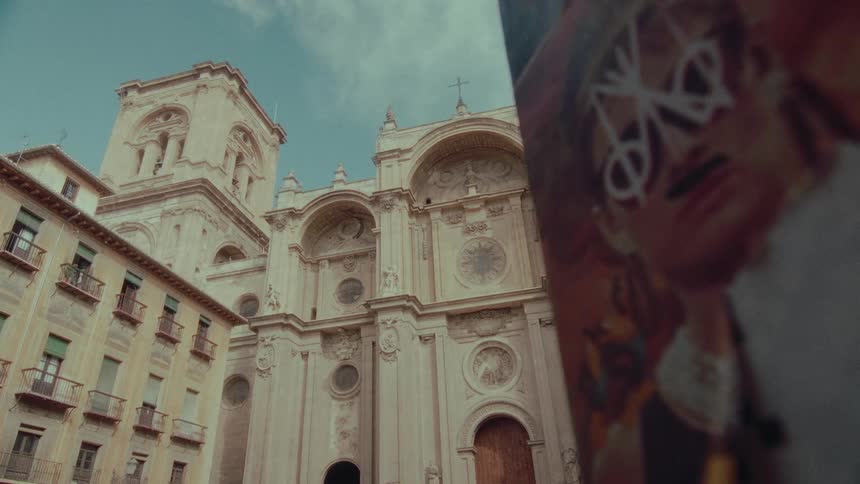 A low-angle view of the majestic Granada Cathedral stands against the sky, with a defaced modern poster in the artistic foreground