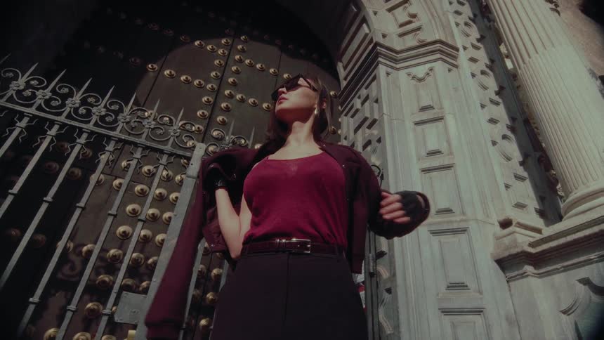 A cool and confident woman in sunglasses and gloves puts her jacket on in slow motion in front of an ornate historical door