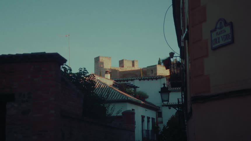 The historic Alhambra palace is visible above the rooftops of a shadowy street in Granada, Spain during a warm sunset