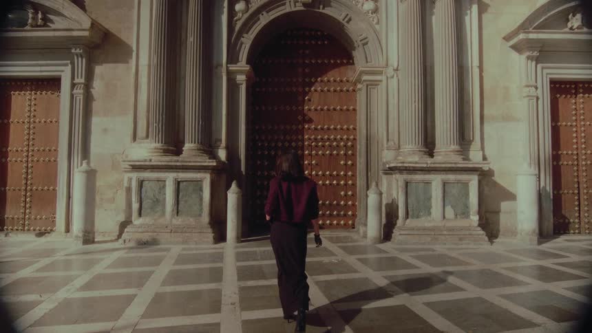 A mysterious and fashionable woman turns and walks towards the grand doors of a sunlit historic building
