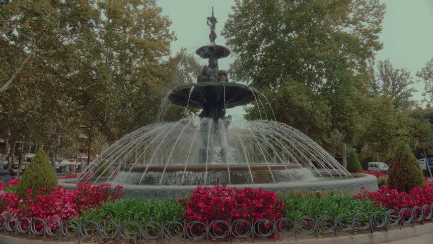 A beautiful ornate water fountain sprays water in a city park as the camera slowly pulls back to reveal vibrant red flowers