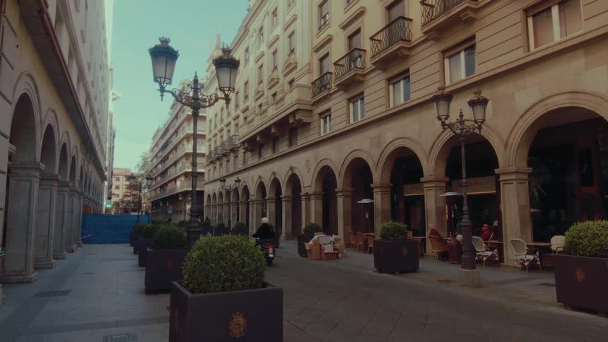 A person rides a scooter down a classic European pedestrian street lined with arched buildings and ornate lampposts
