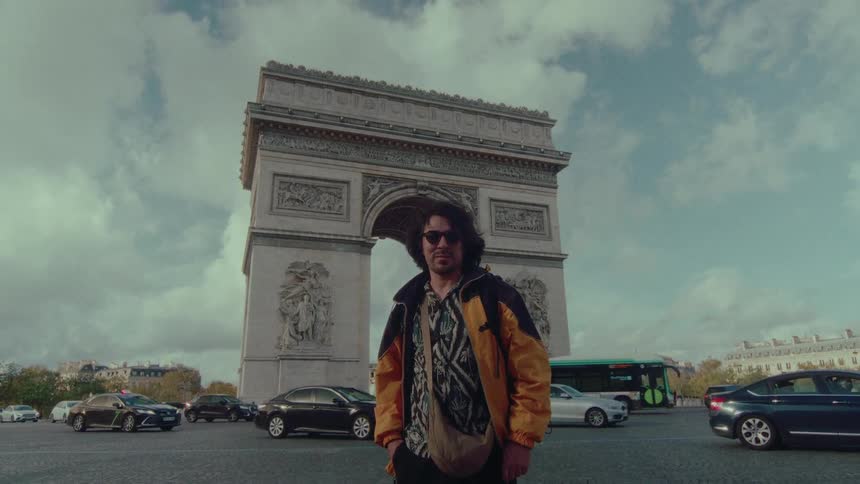 A long-haired man in a yellow jacket stands proudly in front of the Arc de Triomphe as traffic circles around him in Paris, France