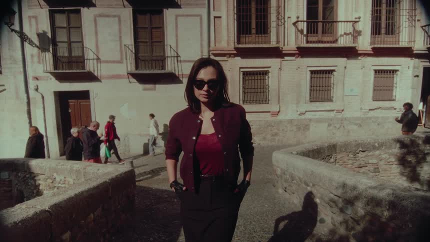 Filmed in slow motion, a mysterious woman in sunglasses and a bomber jacket turns around and walks confidently toward the camera on an old European stone bridge