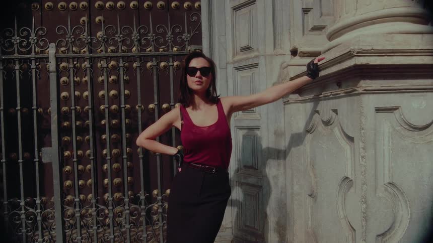 A stylish and mysterious woman in sunglasses and fingerless gloves poses against a historic building's stone column and ornate gate