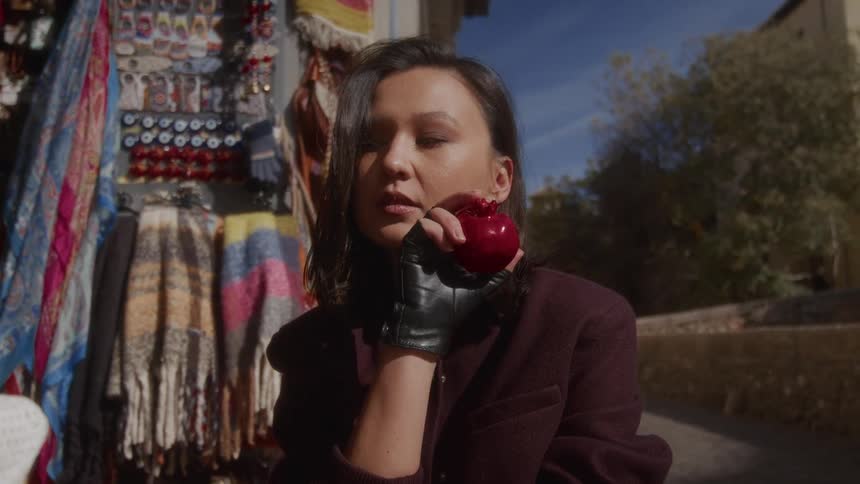 A stylish woman in black leather gloves holds a red pomegranate while posing at an outdoor souvenir market